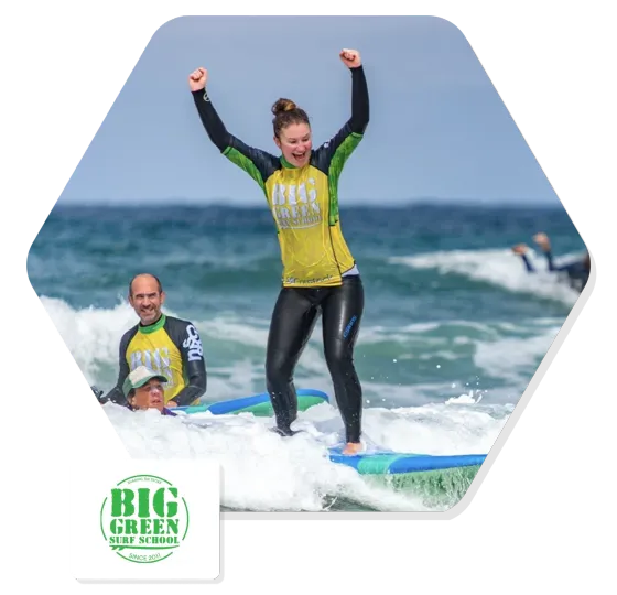 A learner surfer standing up on a wave with both arms raised, cheered on by a Big Green Surf School instructor