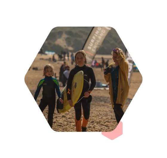 Two young surfers carrying their boards up the beach past a Surfing England flag, with a photographer walking beside them