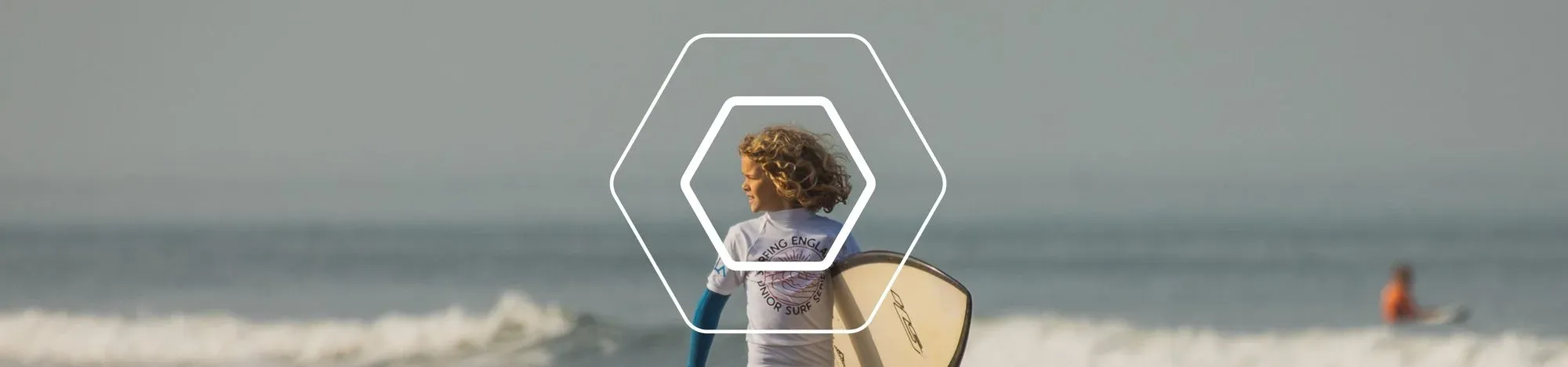 A young surfer carrying a board along the water’s edge, with a surfer riding a wave behind